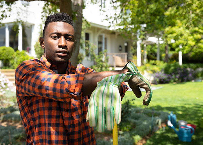 Young man in a plaid shirt putting on gardening gloves outside a newly purchased house on a sunny day, symbolizing homeownership.