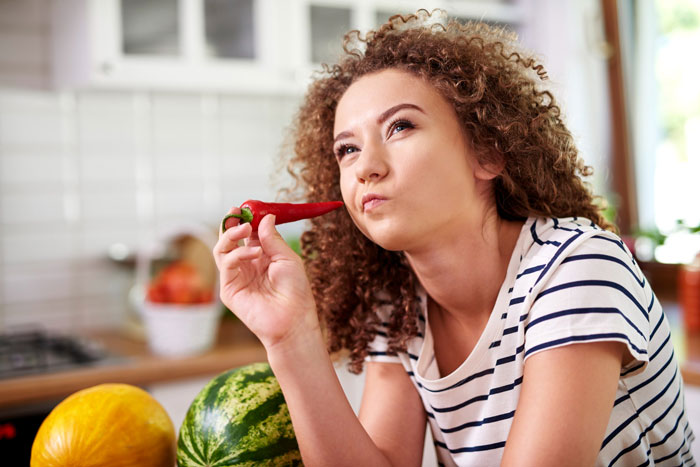 Woman tasting extra-spicy chili pepper in kitchen, experiencing fiery mouth sensation from spicy lunch regret.