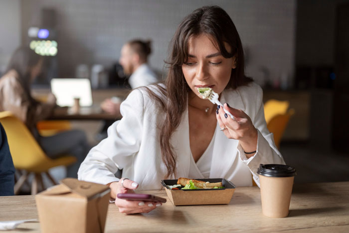 Woman eating spicy lunch at office, holding phone, surrounded by takeaway food containers, expressing regret and discomfort.