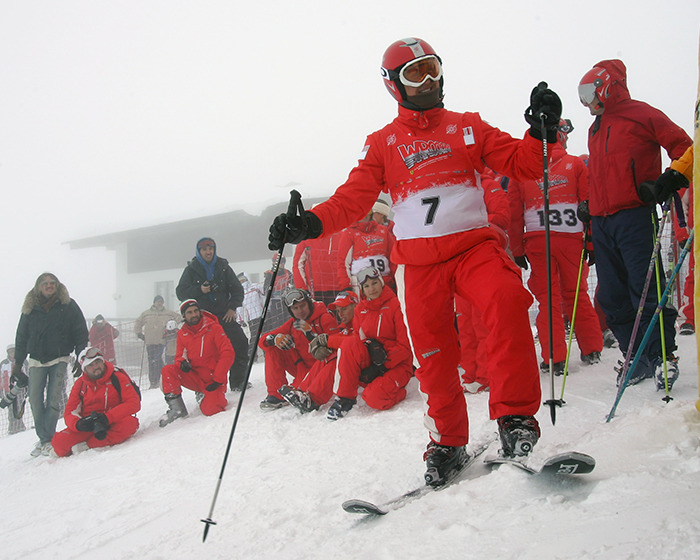 Skier in red gear with number 7 posing on snow while group of people in red ski suits watch in foggy conditions.