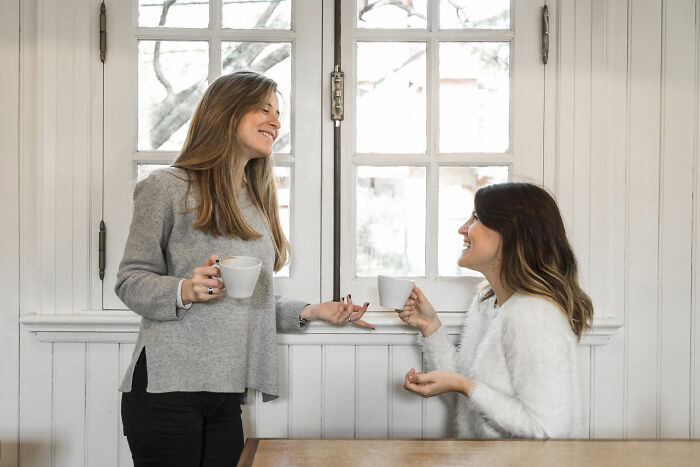 Two women smiling and chatting over coffee in a bright room, depicting a small business owner and friend interaction. Two women smiling and chatting over coffee in a bright room, depicting a small business owner and friend interaction.