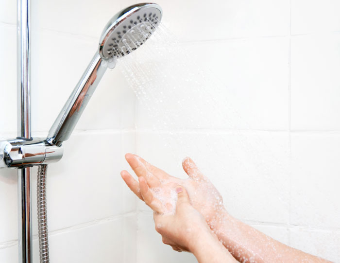 Person washing hands under running water from a modern showerhead in a white tiled bathroom setting.