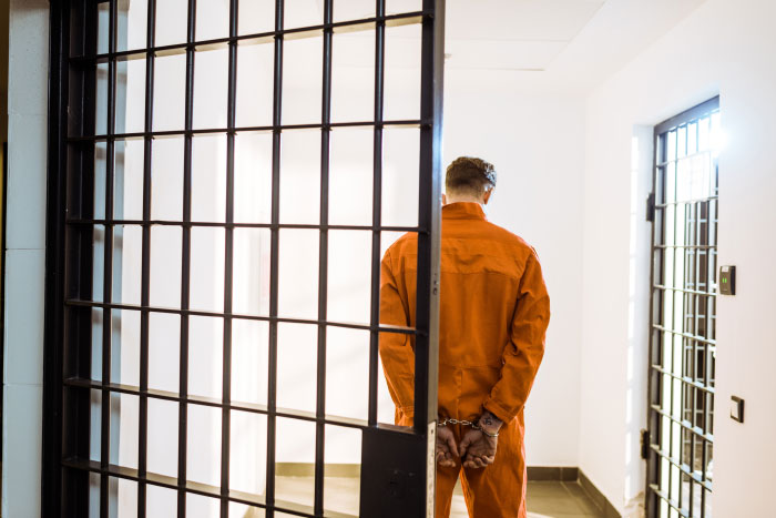 Man in orange prison jumpsuit with handcuffs standing behind jail bars in a bright corridor, symbolizing lying and jail consequences.