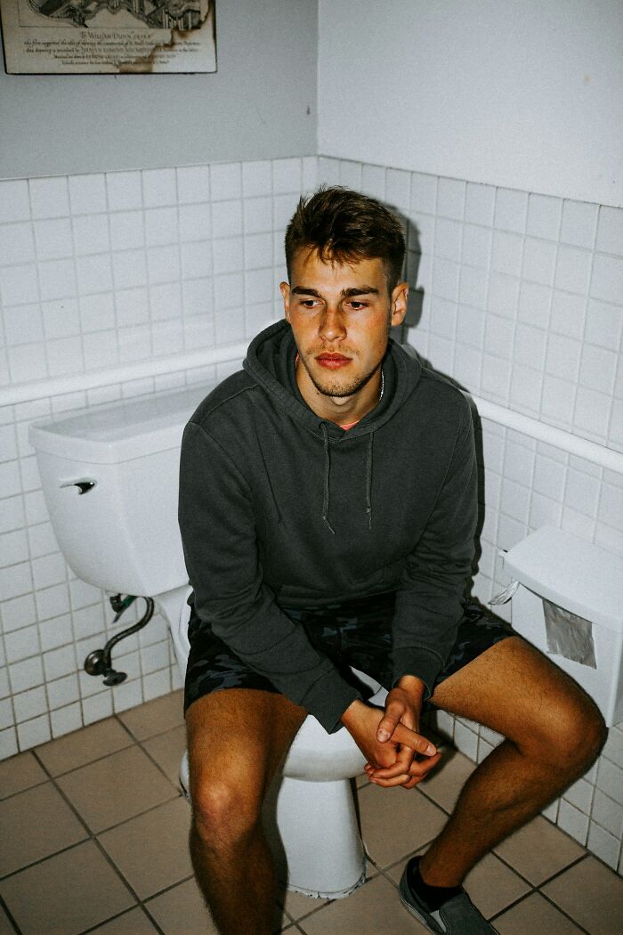 Young man in casual clothes sitting on a toilet looking worried, illustrating accidental texts causing panic situations.