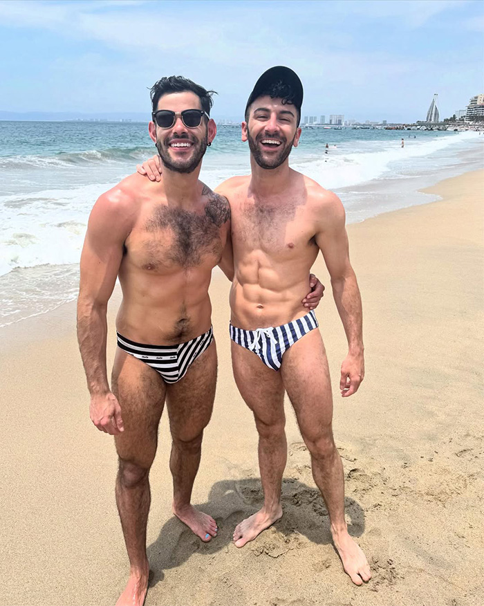 Gay couple smiling arm in arm on sandy beach wearing striped swim briefs, ocean and skyline in background Gay couple smiling arm in arm on sandy beach wearing striped swim briefs, ocean and skyline in background