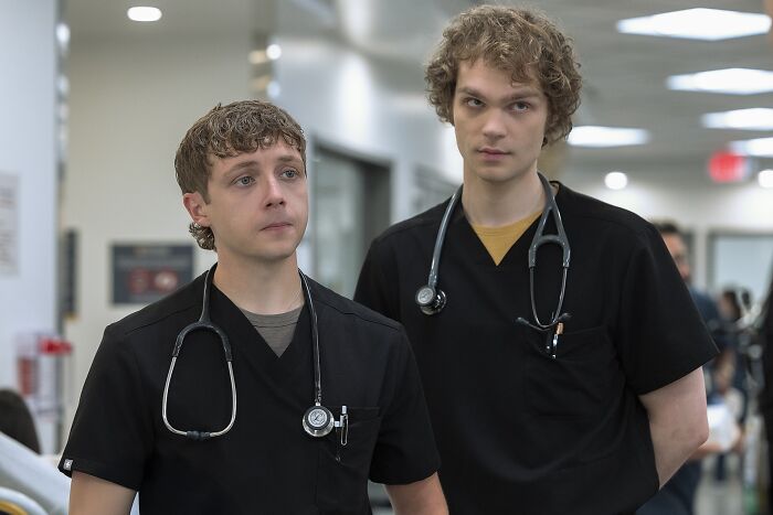 Two young male medical interns wearing black scrubs and stethoscopes in a hospital hallway, Insufferable Pitt fans discussion.