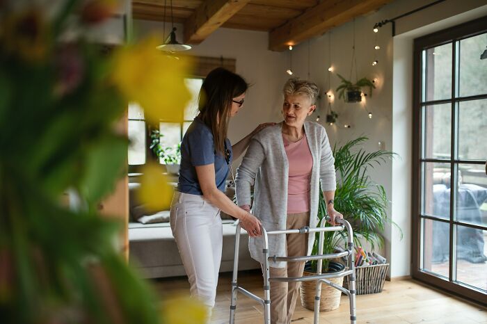 A young woman helping an elderly woman with a walker indoors, highlighting cultural differences and normal behaviors.