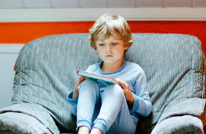 Young child in pajamas sitting on a couch, deeply focused on reading a book, showcasing surprising intelligence.