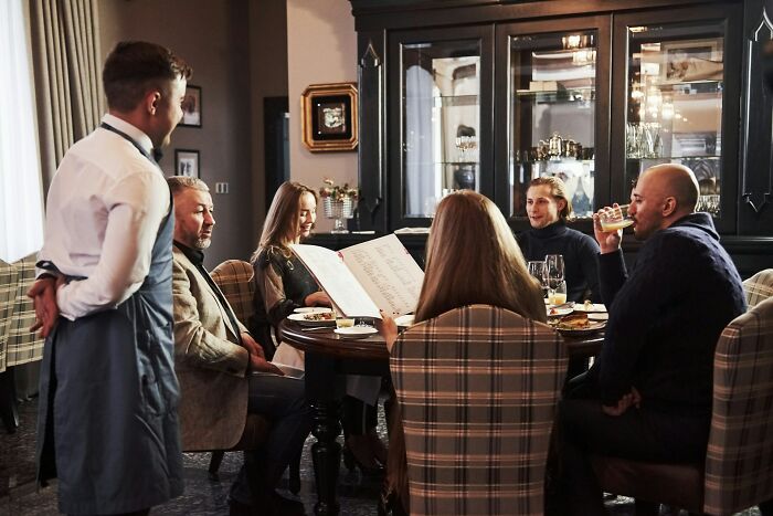Group of people dining and interacting with a waiter, showcasing normal practices in Europe that the US may find weird.