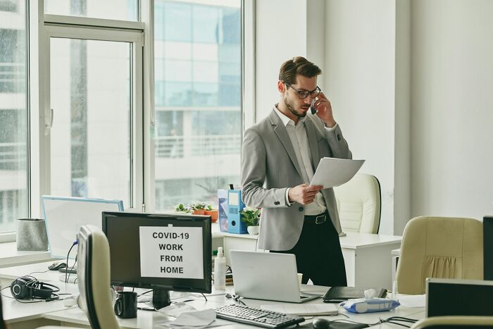 Man in office reading papers and talking on phone, with a computer screen showing work from home during COVID-19 and shipping containers concept.