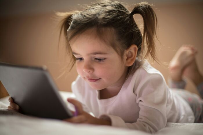 Young girl lying on bed using tablet, illustrating concerns related to pressing issues plaguing America today.