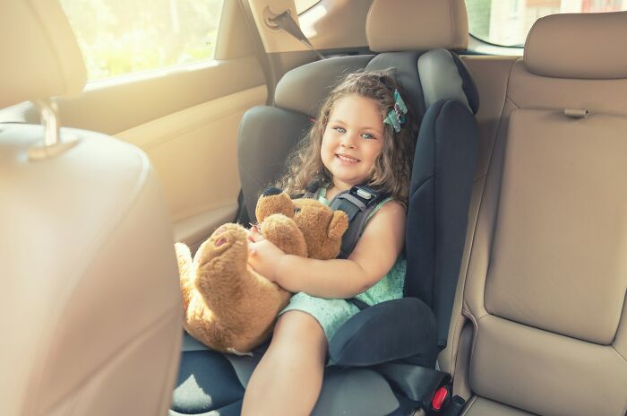 Young girl smiling in car seat holding teddy bear, showcasing surprising intelligence in an everyday moment.