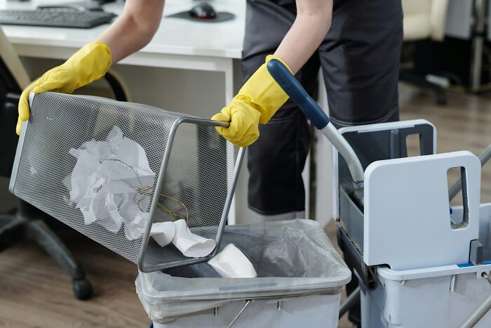 Person wearing yellow gloves emptying office trash can near cleaning cart showing worst coworkers at work environment
