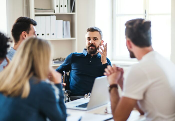 Man in wheelchair sharing coworker stories during a meeting with colleagues in a bright office setting.