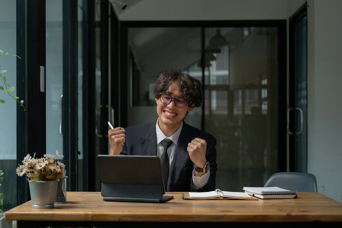 Young man in glasses celebrating success working on a tablet in a modern office, illustrating adult goals achievement.