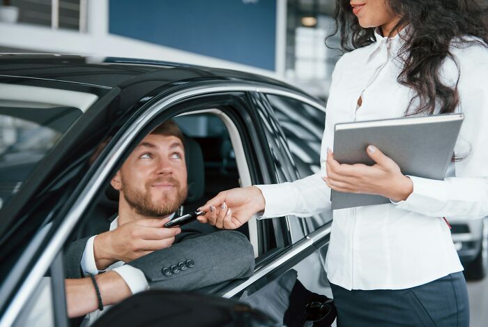 Man sitting in new car receiving keys from female car dealership employee holding a tablet in showroom.