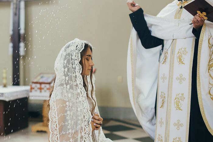 A woman in a lace veil kneeling as a priest sprinkles holy water, symbolizing former cult members' awakening moments.