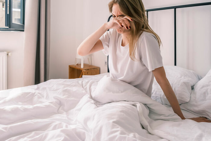 Woman sitting on bed, wiping her eyes, expressing distress related to food shopping triggering emotional response.