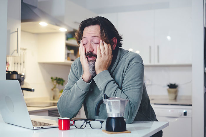 Frustrated man in kitchen sitting at table with laptop and coffee, symbolizing gifted kids not living up to potential.