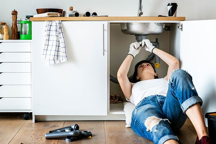 Woman fixing plumbing under kitchen sink, wearing gloves and casual clothes, showing independence and strength.