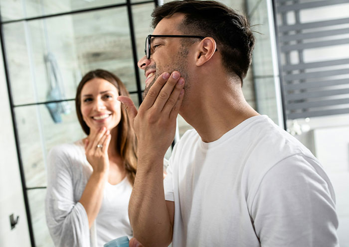 Man shaving in bathroom mirror while woman watches, illustrating woman demands swimmer boyfriend look like her friend&rsquo;s partner.