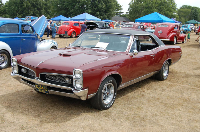 Classic red Pontiac car at an outdoor show, illustrating issues with BF&rsquo;s car during GF&rsquo;s parents visit.