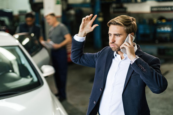 Frustrated man talking on phone near car in auto repair shop, highlighting problems with boyfriend&rsquo;s car and parents&rsquo; visit.