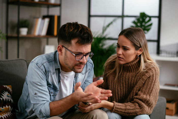 Couple having a tense conversation on couch, woman demanding boyfriend man up and console her over mother-in-law loss.