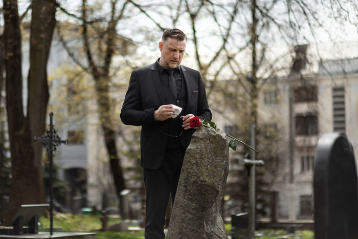 Man grieving at tombstone in cemetery holding rose, reflecting on loss and emotional demands during mourning.