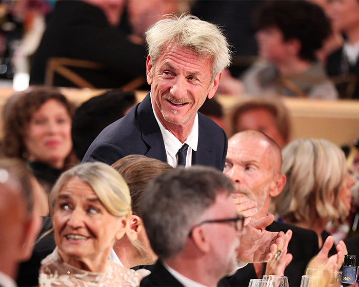 Man in suit smiling among seated guests at 2026 Golden Globes event with visible chaos and distracted attendees around.