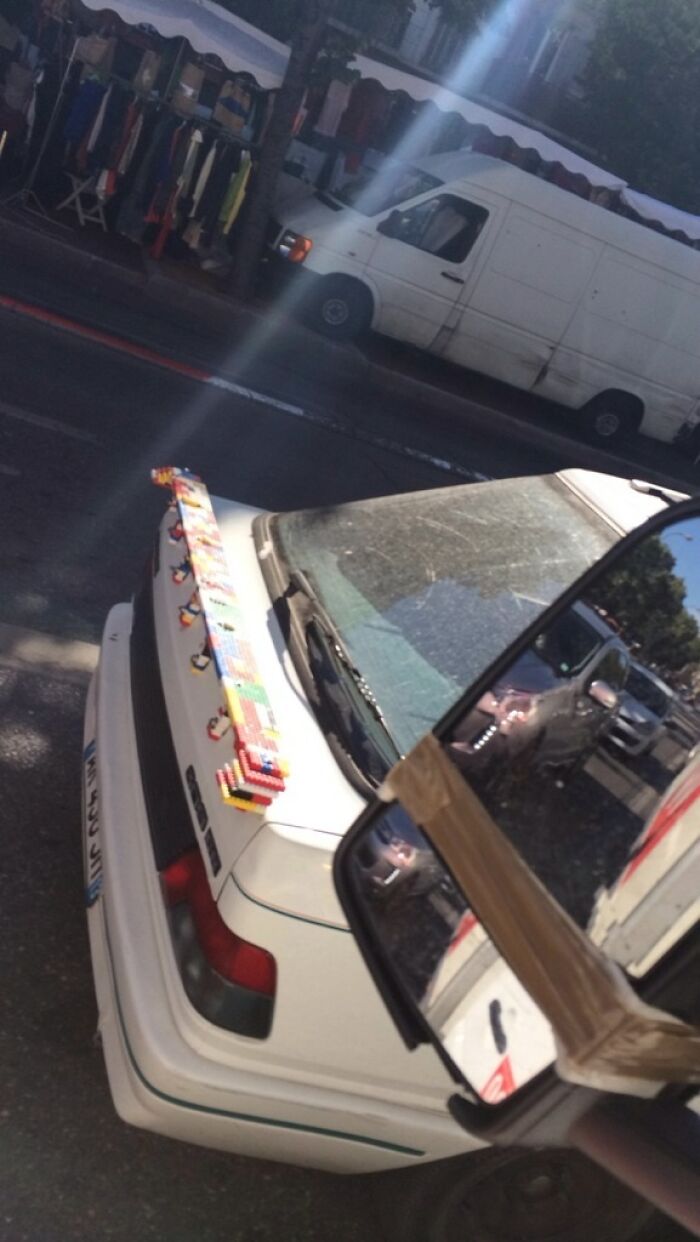 White car with a homemade DIY spoiler made from colorful Lego blocks parked on a street near a white van.