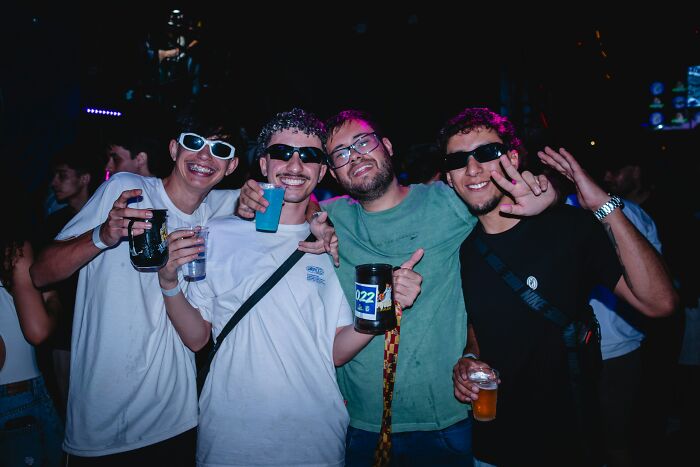 Group of young men wearing sunglasses and holding drinks, enjoying a lively nightclub party atmosphere.