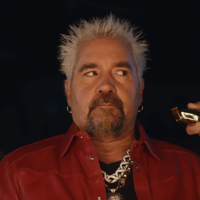Guy Fieri with spiky blonde hair, wearing a red leather jacket and chain necklace in a dimly lit setting.