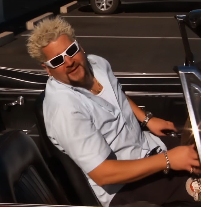 Guy Fieri sitting in a car wearing white sunglasses and a light blue shirt, showing his spiky blonde hair style.