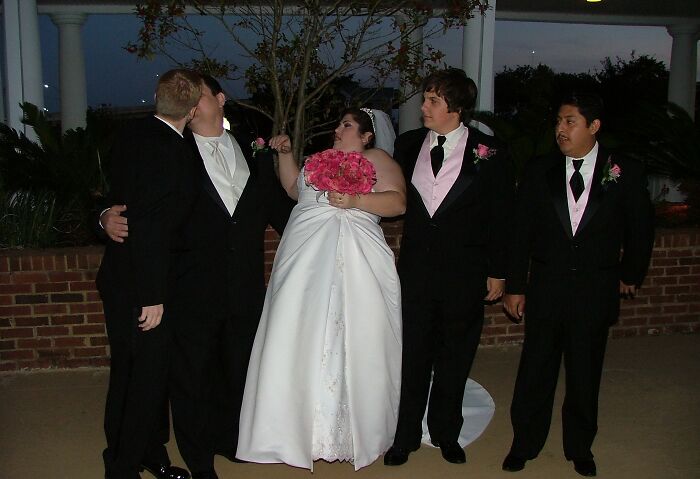 Bride holding bouquet looks surprised as two groomsmen kiss while other groomsmen watch in awkward wedding photo.