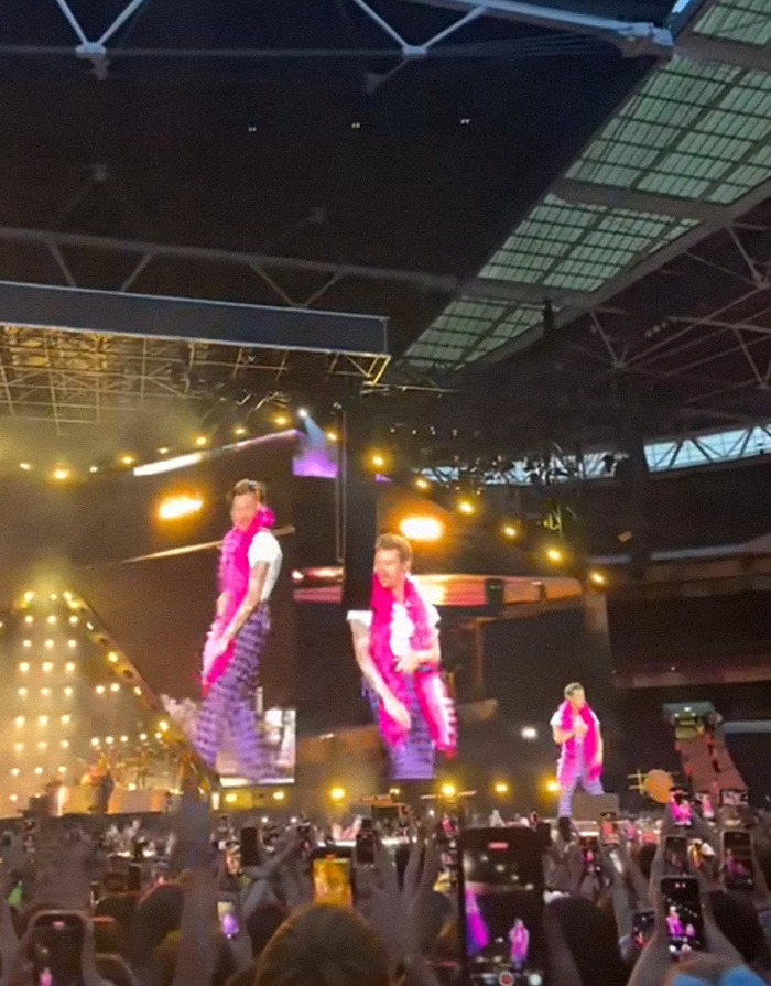 Harry Styles performing on stage wearing a pink feather boa during his new tour concert with fans recording.