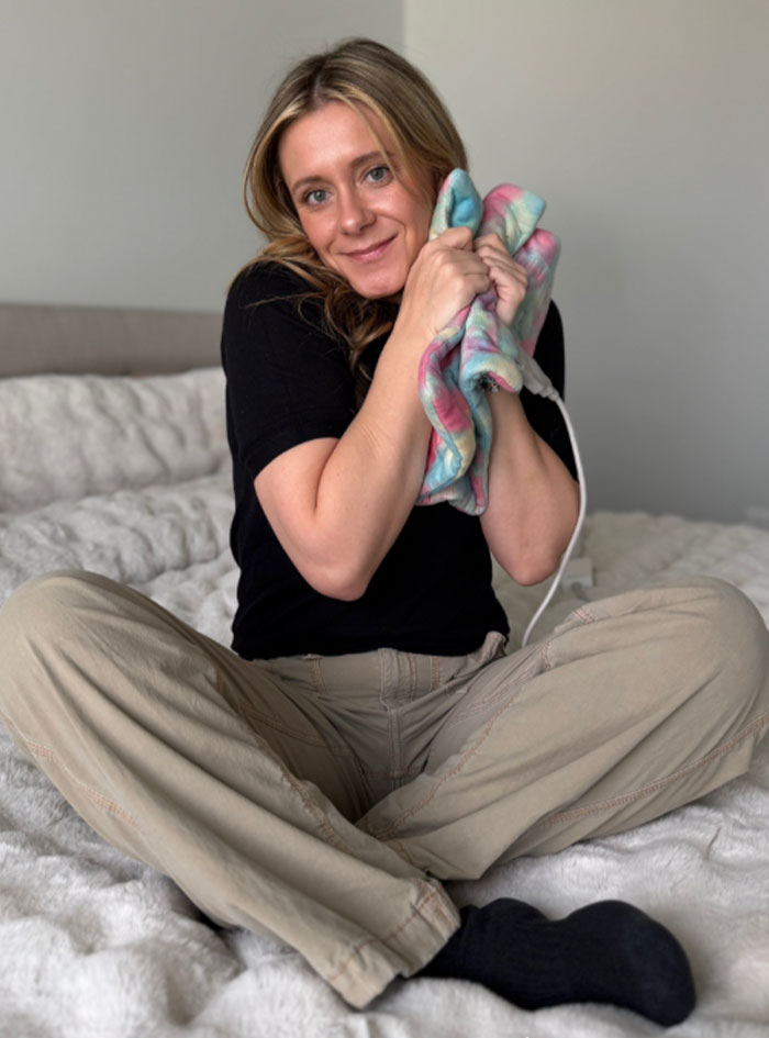 Woman sitting on bed holding a heating pad close to her face, demonstrating use related to toasted skin syndrome.
