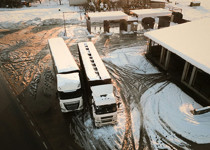 Two snow-covered trucks parked near a building with snowy and muddy tire tracks on the ground at sunset.