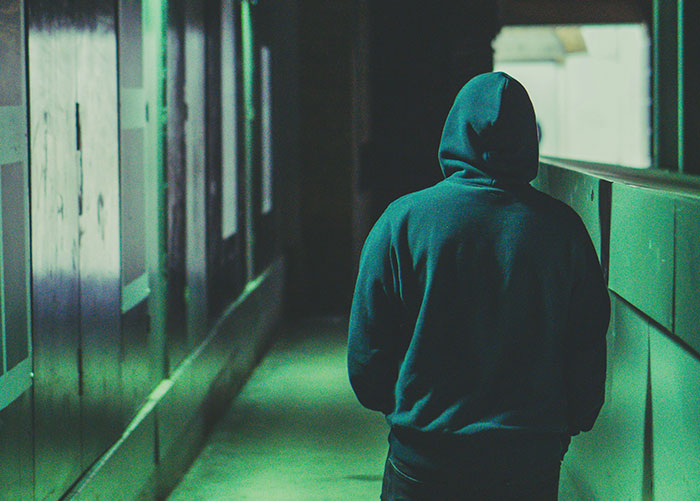 Person wearing a dark hoodie walking alone in a dimly lit alley reflecting experiences of unhoused people on the streets.