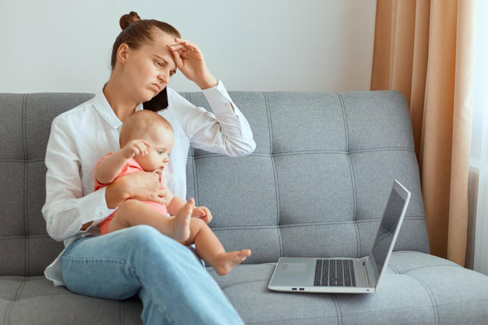 Stressed woman holding baby on couch, looking upset while working on laptop, illustrating child-loathing and nanny struggles.