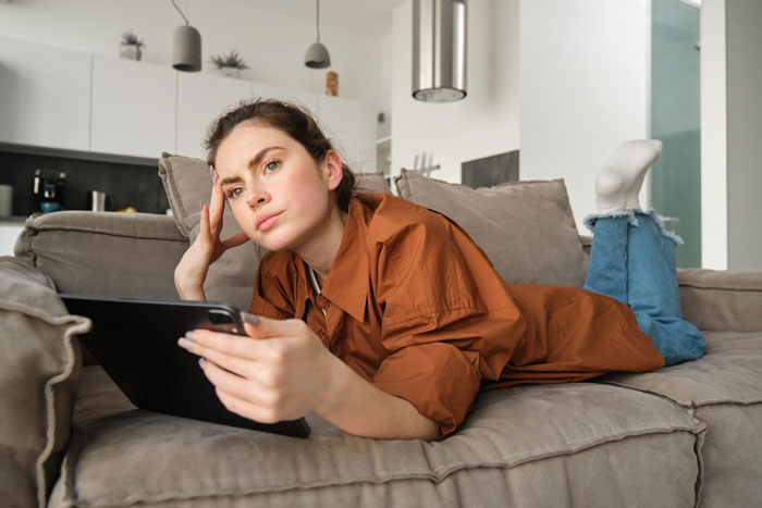 Young woman lying on couch, looking thoughtful and concerned while holding a tablet about inherited home issues. Young woman lying on couch, looking thoughtful and concerned while holding a tablet about inherited home issues.