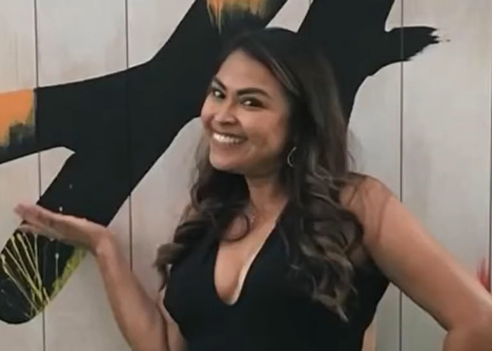 Smiling woman with long hair posing indoors with an abstract black and orange wall art behind her, representing teacher&rsquo;s life story.