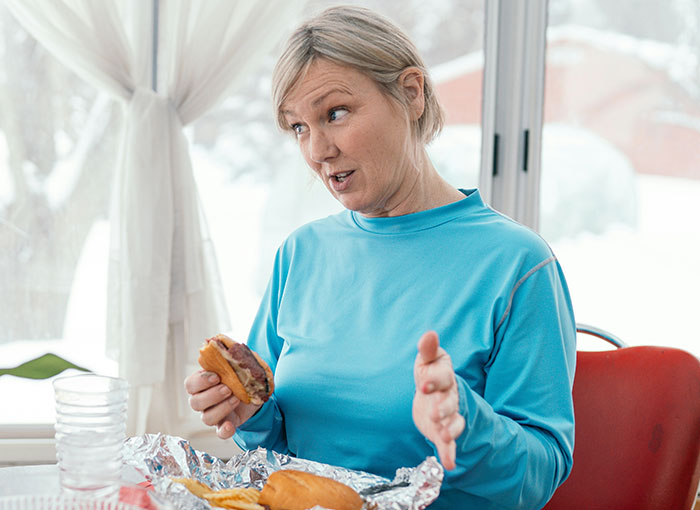 Woman in a blue shirt eating a sandwich while discussing a fun fact with a child and his dad nearby.