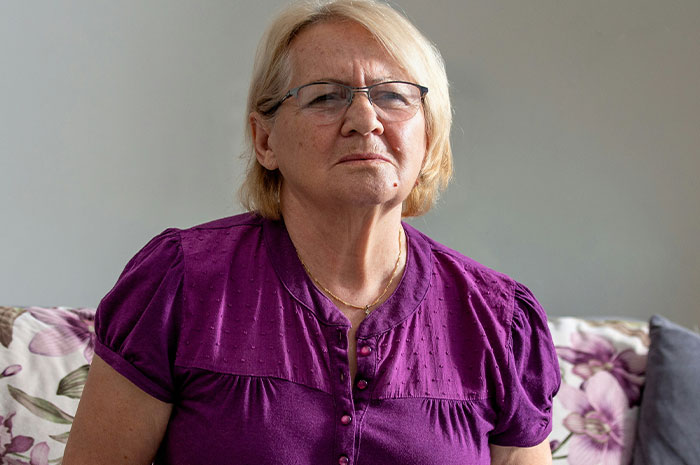 Elderly woman in glasses and purple blouse sitting on floral couch, related to husband parents coming honeymoon surprise.
