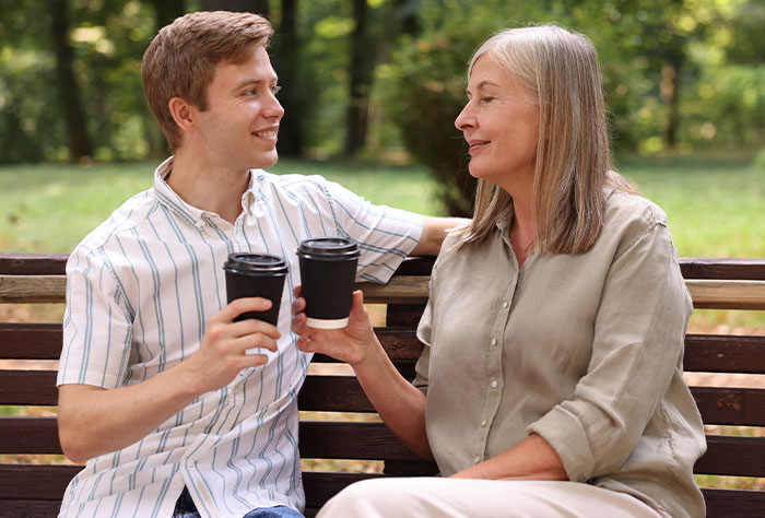 Man and his mother‑in‑law sitting on a bench outdoors, sharing coffee, highlighting family tensions and support dynamics.