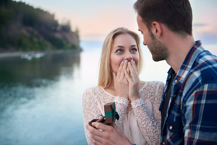 Couple by the water with man proposing and woman reacting with surprise, illustrating a Venmo search unraveling a fabricated past.
