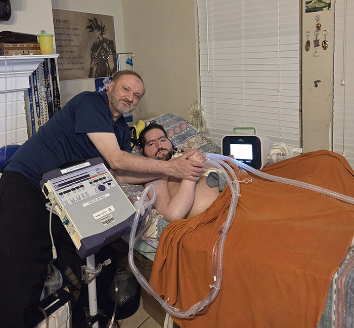A father embraces his bedridden son connected to medical equipment in a room, highlighting detained father funeral issue.