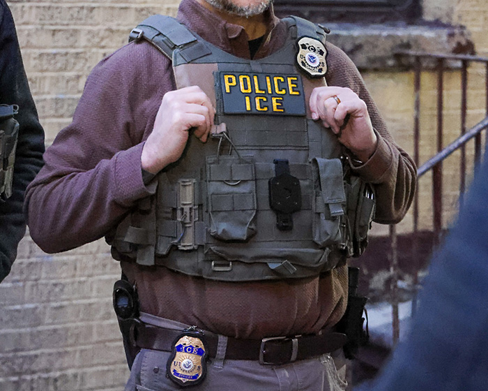 ICE officer wearing tactical vest with police and ICE patches during an enforcement operation outside a brick building.