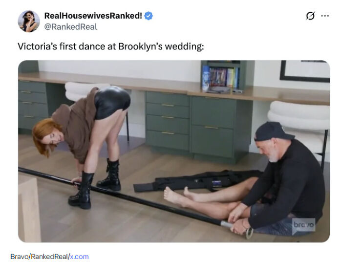 Woman in brown sweater and black boots dancing playfully indoors while a man in black hat and shirt holds a pole for support