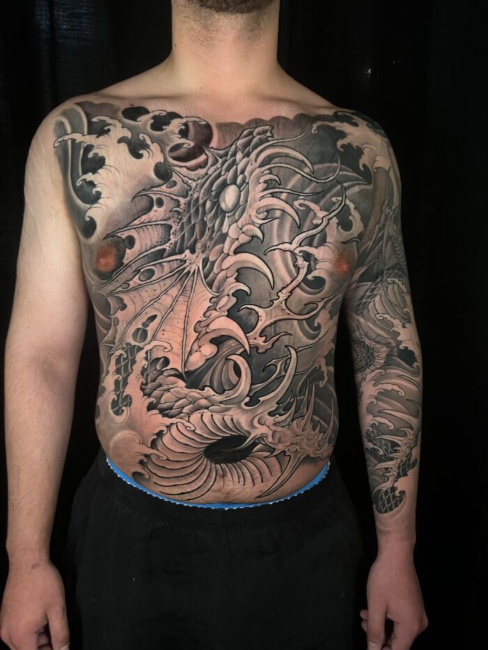 Intricate dragon tattoo covering chest and arm, showcasing detailed shading and design in a realistic style.